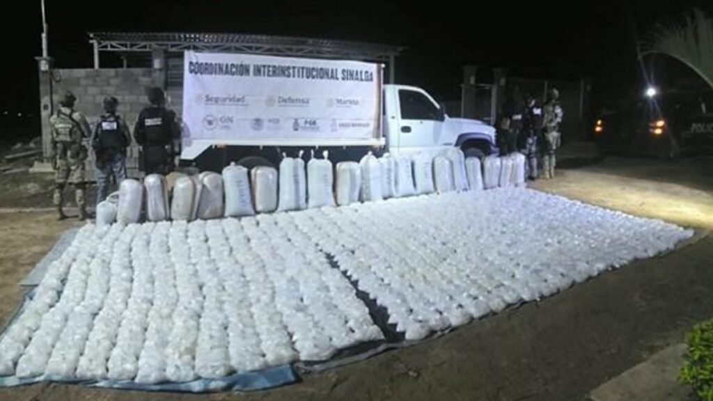 Hundreds of clear plastic packages of methamphetamine laid out on a tarp at night in front of a white pickup truck and a 'Coordinación Interinstitucional Sinaloa' banner, with soldiers and police standing behind.