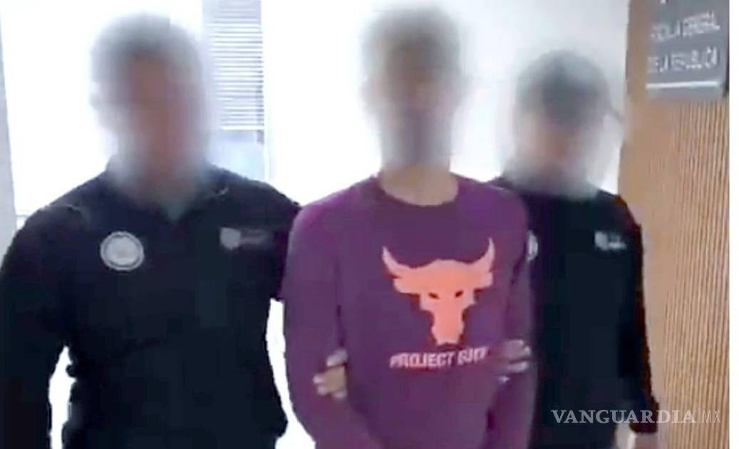 José Ángel Rivera Zazueta, face blurred, in a maroon “Project Rock” long-sleeve shirt, held by the arms by two agents in black uniforms in a hallway beside a Fiscalía General de la República sign