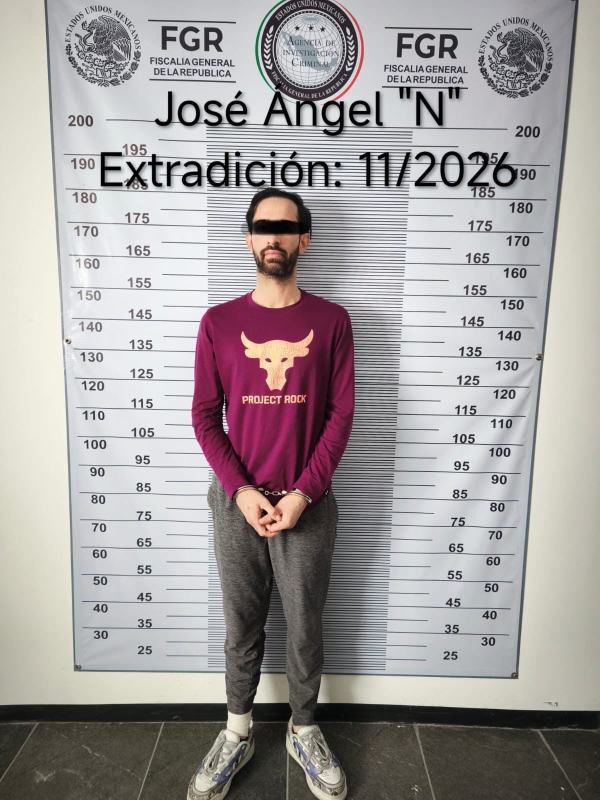 José Ángel Rivera Zazueta, eyes blacked out, handcuffed in a maroon long-sleeve shirt and gray sweatpants, standing before an FGR height chart labeled “José Ángel N, Extradición: 11/2026”