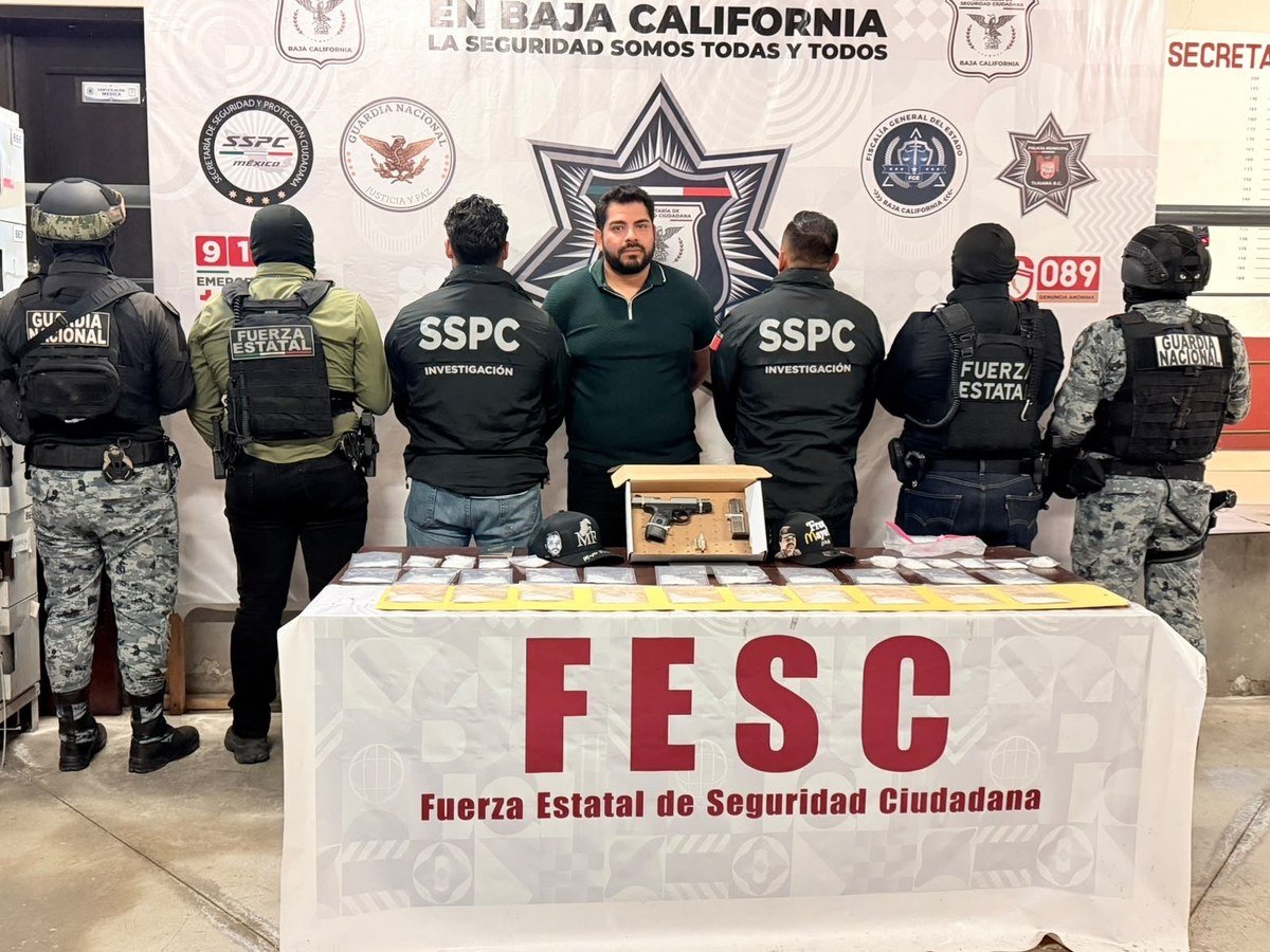 Luis Adán Torales Castillo, alias 'El 500,' stands between Baja California state police and federal SSPC agents behind a FESC table displaying a 9mm pistol, small bags of drugs and two caps