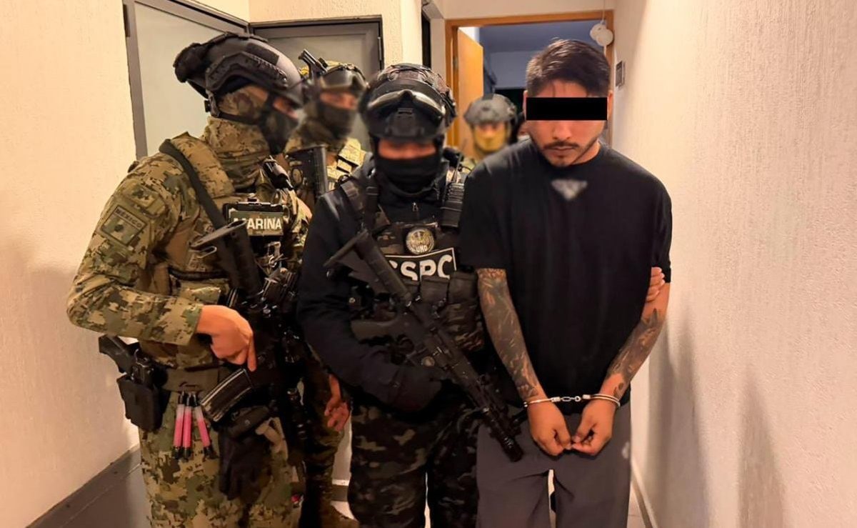 A handcuffed man in a black T-shirt, his eyes covered by a black bar, is led down a hallway by a Mexican Navy marine and an SSPC federal agent in tactical gear.