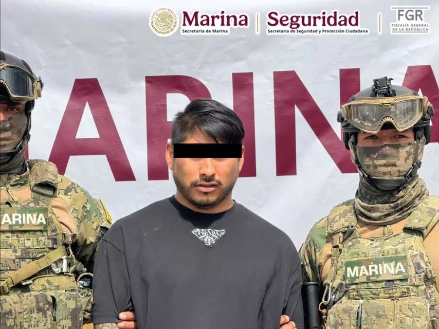 A man in a black T-shirt, his eyes covered by a black bar, stands handcuffed between two masked Mexican Navy marines in front of a banner reading Marina.