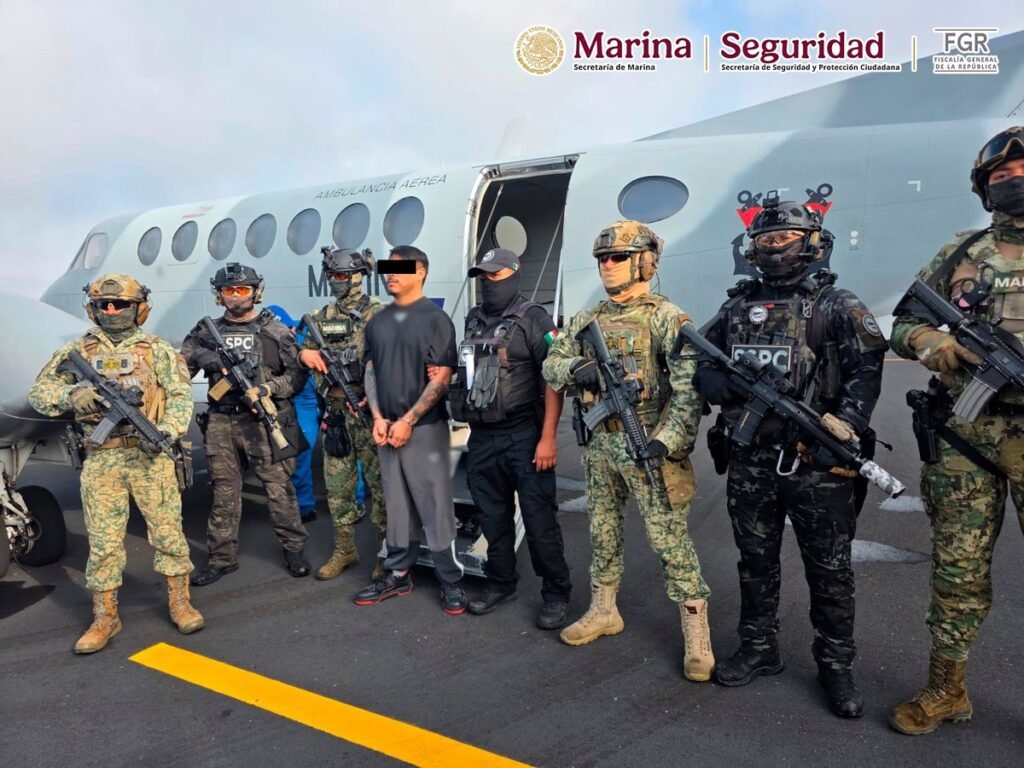 A handcuffed man in a black T-shirt, his eyes covered by a black bar, stands on a runway in front of a Mexican Navy air-ambulance plane, flanked by armed Navy marines and SSPC federal agents.