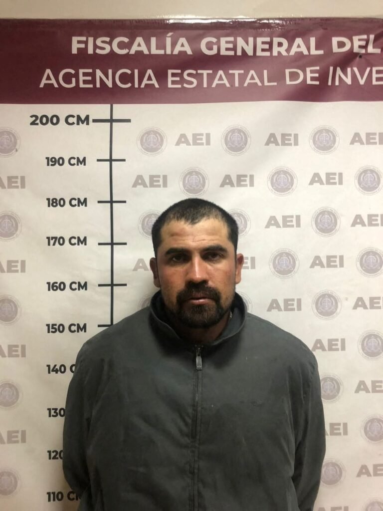 Booking photo of Jesús Gerardo, alias El Kekas, a bearded man in a dark gray zip jacket, in front of a Baja California state investigations agency height chart