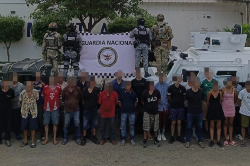 Masked federal agents stand behind a Guardia Nacional banner over a line of 28 detainees whose faces are blurred, in Mazatlán, Sinaloa