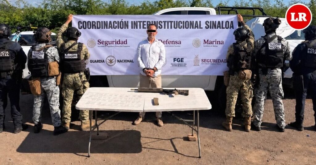 A man in a white shirt, face blurred, stands handcuffed before a 'Coordinación Interinstitucional Sinaloa' banner, flanked by soldiers, National Guard and state police, with a rifle on a table in front of him