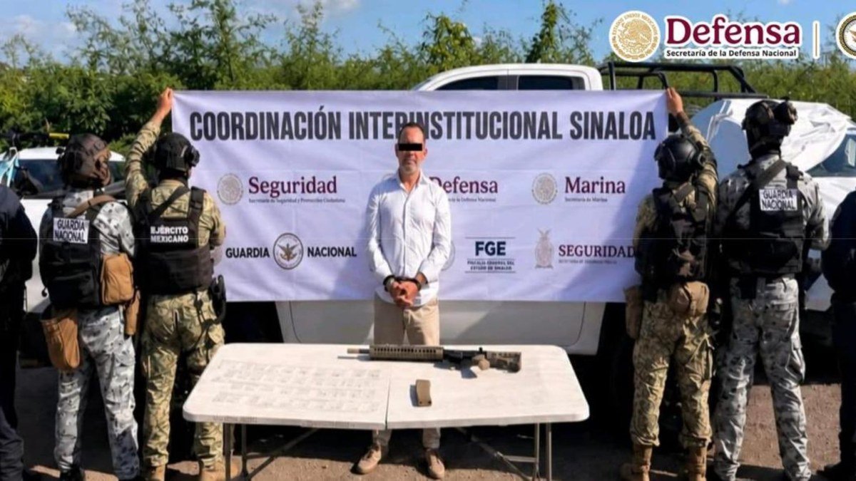A man in a white shirt, face blurred, stands handcuffed in front of a 'Coordinación Interinstitucional Sinaloa' banner held by soldiers and National Guard troops, with a rifle and a magazine on a table before him