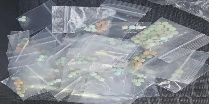 Dozens of small clear zip-lock bags holding small green and orange pills spread on a dark surface