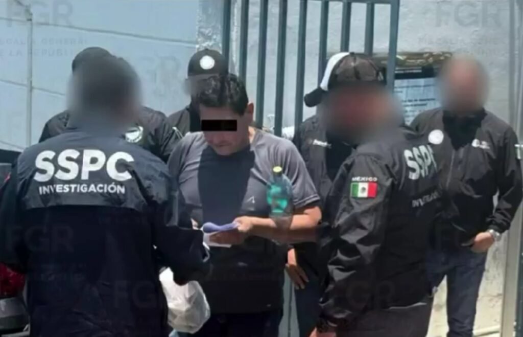 A man in a gray T-shirt, his eyes covered by a black bar, holds a document while surrounded by agents in black SSPC Investigación jackets