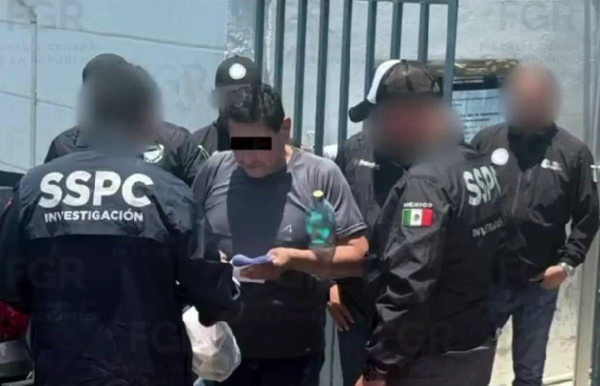 A man in a gray T-shirt, his eyes covered by a black bar, holds a document while surrounded by agents in black SSPC Investigación jackets