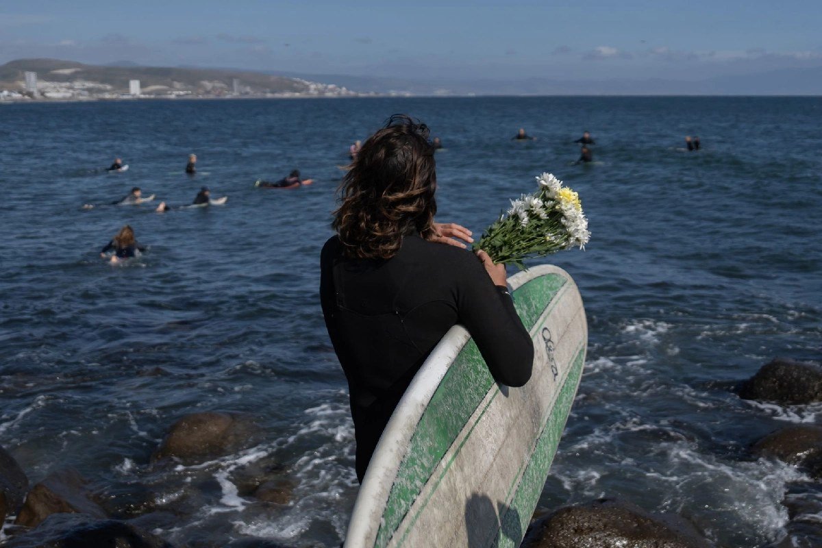 A surfer holding a surfboard and white flowers stands on rocks facing the ocean as other surfers paddle out in Ensenada