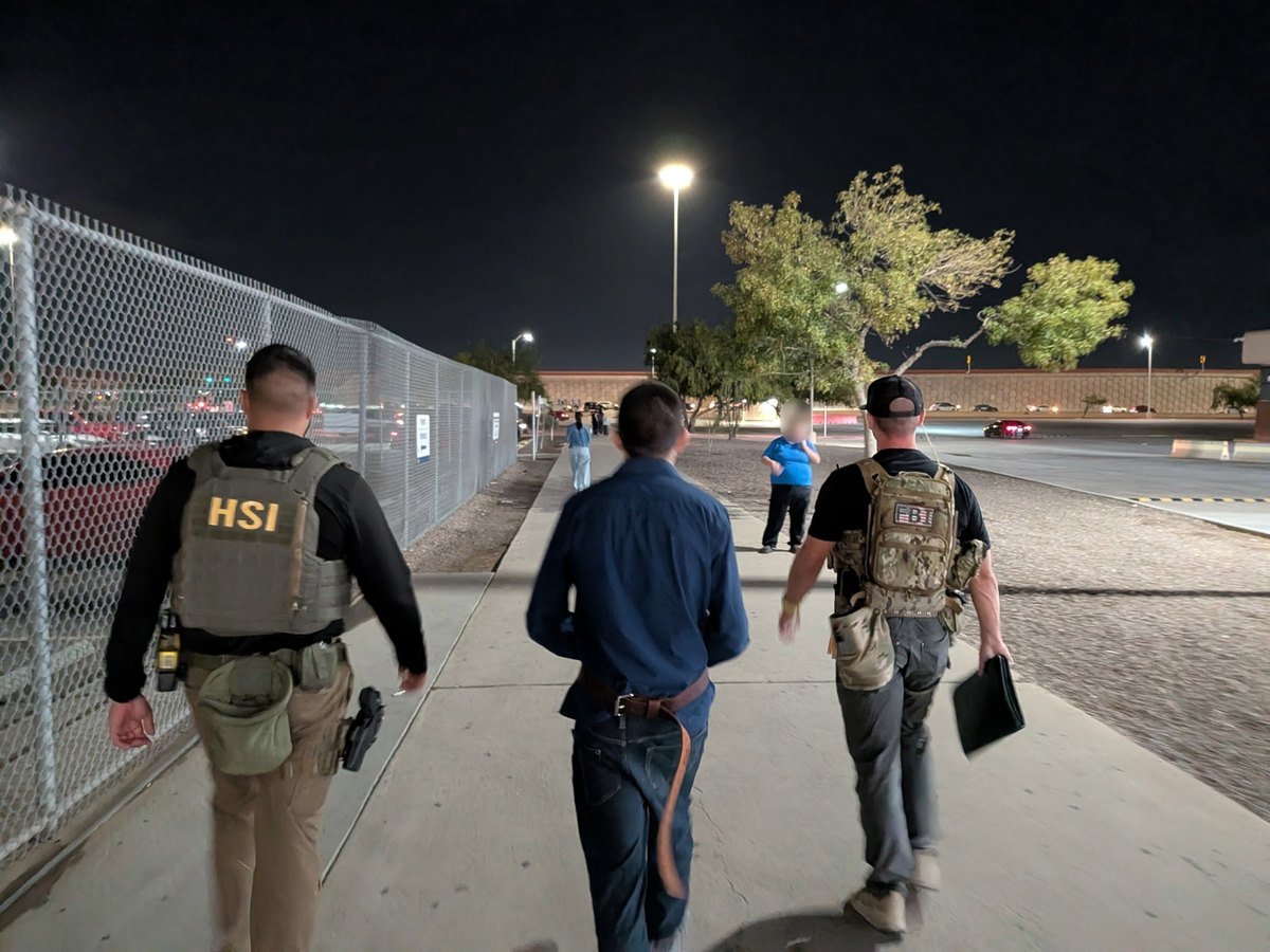 At night, an HSI agent and an FBI agent walk a man in a blue shirt along a fenced sidewalk