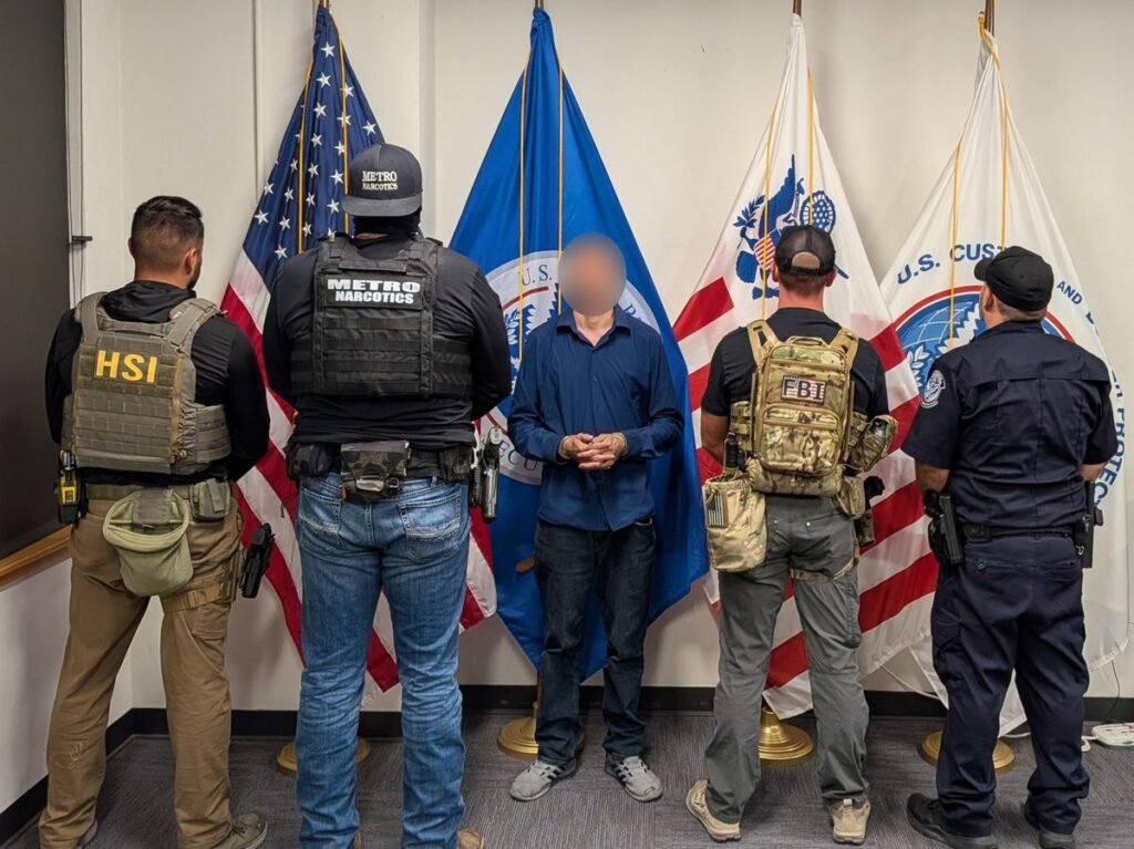 Felix Anthony Salazar III, face blurred, in a blue shirt with hands cuffed in front, standing among HSI, Metro Narcotics, FBI and CBP officers before U.S. and Homeland Security flags