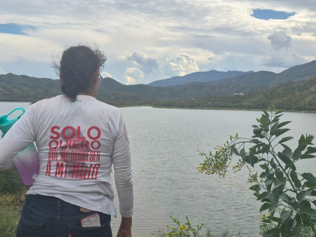 A woman seen from behind, wearing a white shirt with red lettering asking for her son back, looks out over a large reservoir ringed by green hills