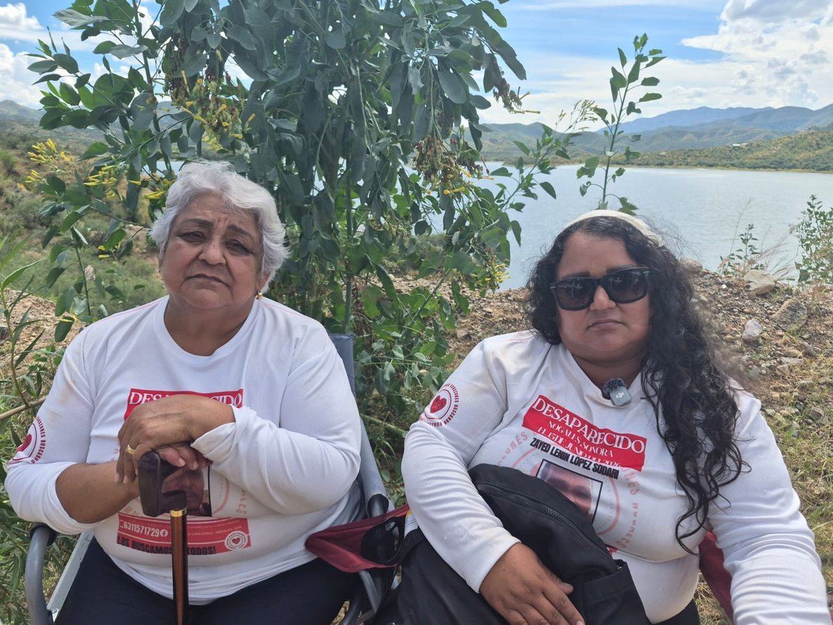 An older woman with a cane and a younger woman in sunglasses sit by a reservoir, both in white shirts printed with a missing man's name and photo