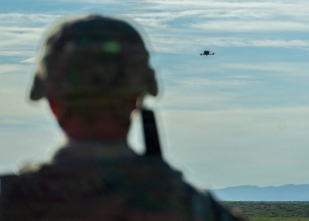 A soldier in a helmet, out of focus in the foreground, watches a small drone flying over desert scrub and distant mountains