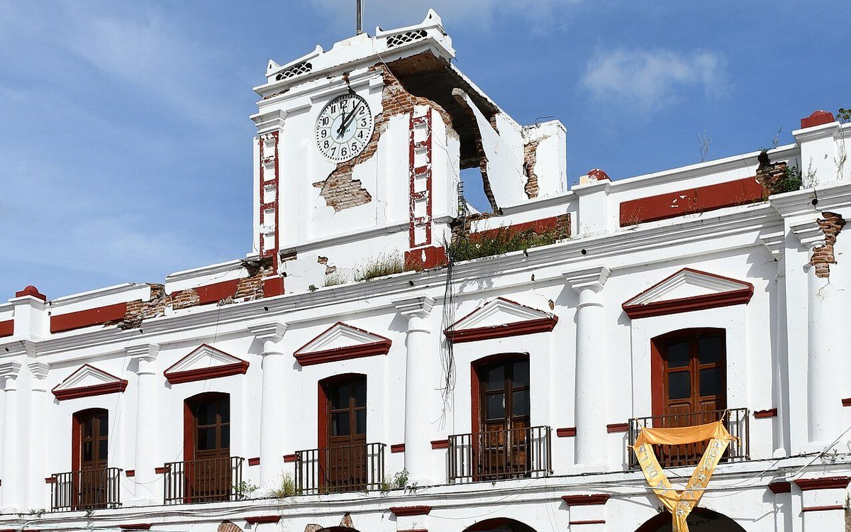 White colonial-style municipal palace in Juchitán, Oaxaca, with red trim, arched balconies and a clock tower showing cracked and missing masonry