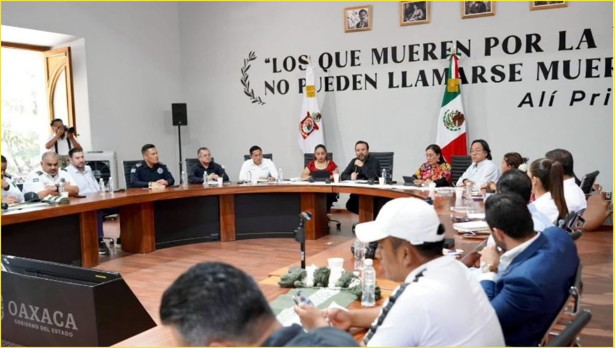 Officials in white and dark shirts, some in police uniforms, sit around a curved conference table under Mexican and Oaxaca flags during a government meeting.