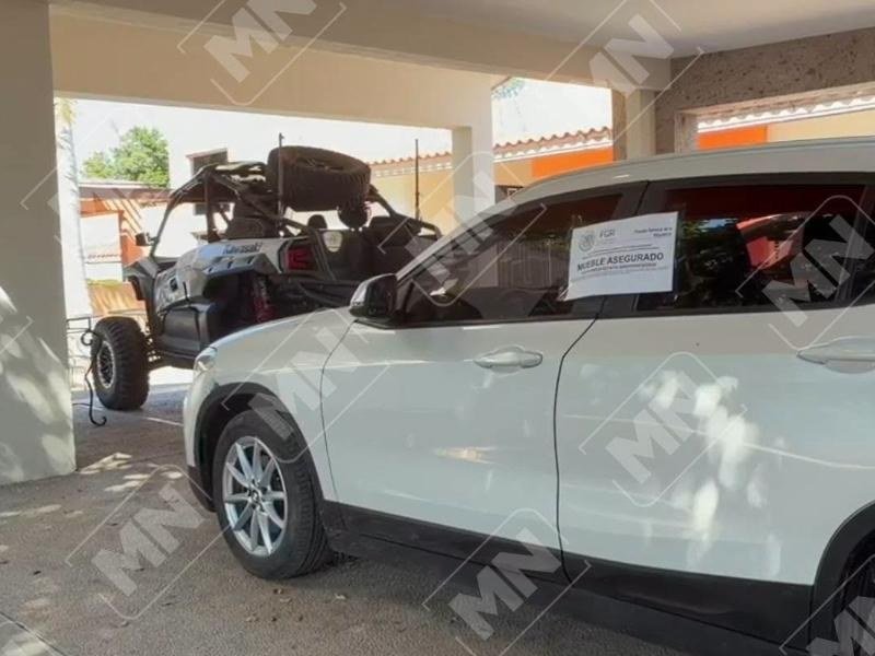 A white SUV with a federal prosecutors' 'mueble asegurado' seizure notice on its window, parked under a carport next to an off-road buggy
