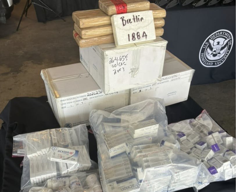 Wrapped bricks labeled with marker sit on cardboard boxes beside bags of boxed medicine on a table in front of a Department of Homeland Security banner