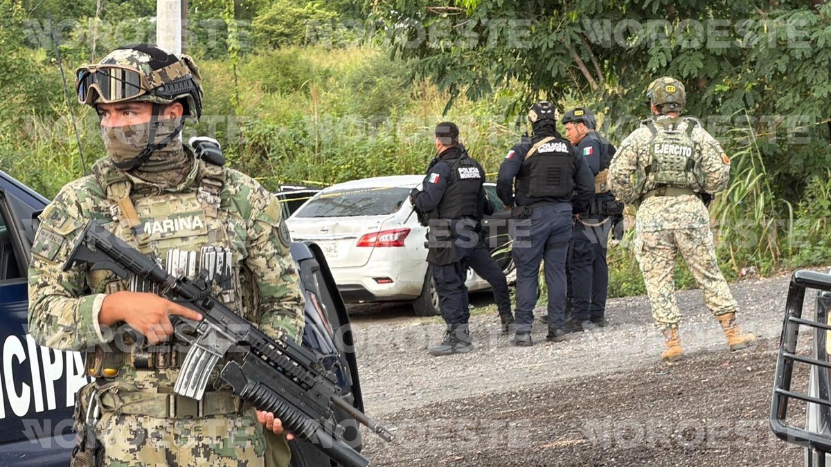 A Mexican Navy marine with a rifle stands guard as municipal police and Army soldiers stand beside a white Nissan sedan left in brush beside the highway near Mazatlán's airport