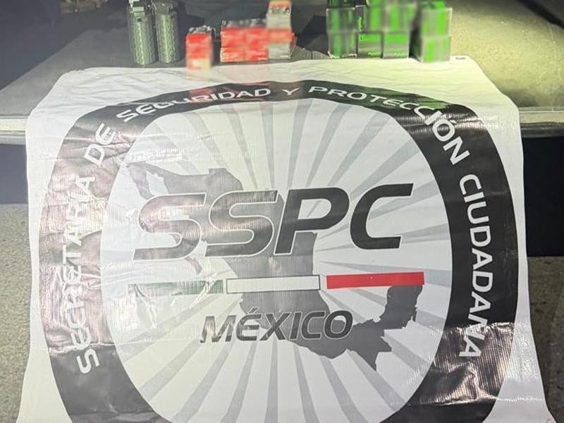 Grenades and boxes of ammunition, partly blurred, displayed on a table behind a white banner bearing the logo of Mexico's Security and Citizen Protection Secretariat (SSPC).