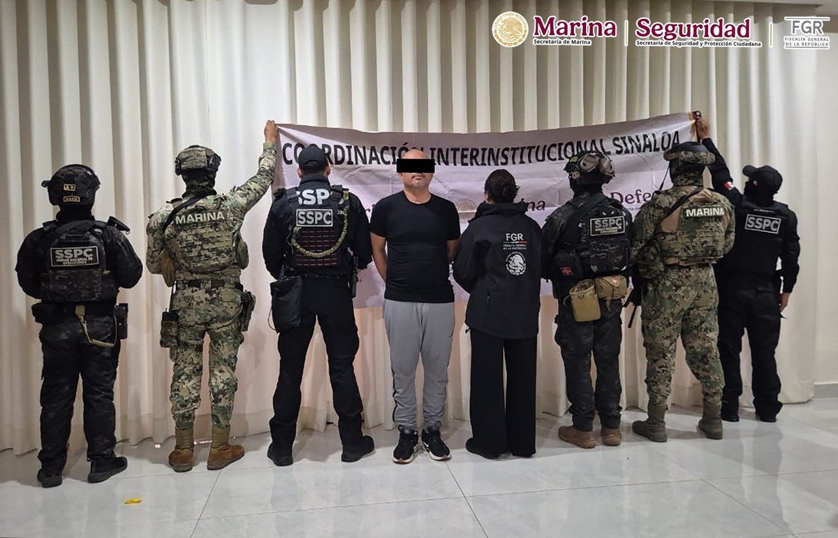 A detained man in a black T-shirt and gray sweatpants, his eyes blacked out, stands between Navy, SSPC and FGR personnel in front of a banner reading Coordinación Interinstitucional Sinaloa