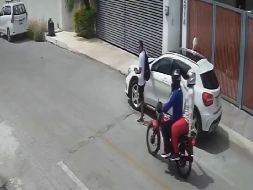 Security-camera frame of two riders on a red motorcycle pulling up beside a white SUV on a residential street in Mérida as a man stands by the car door.