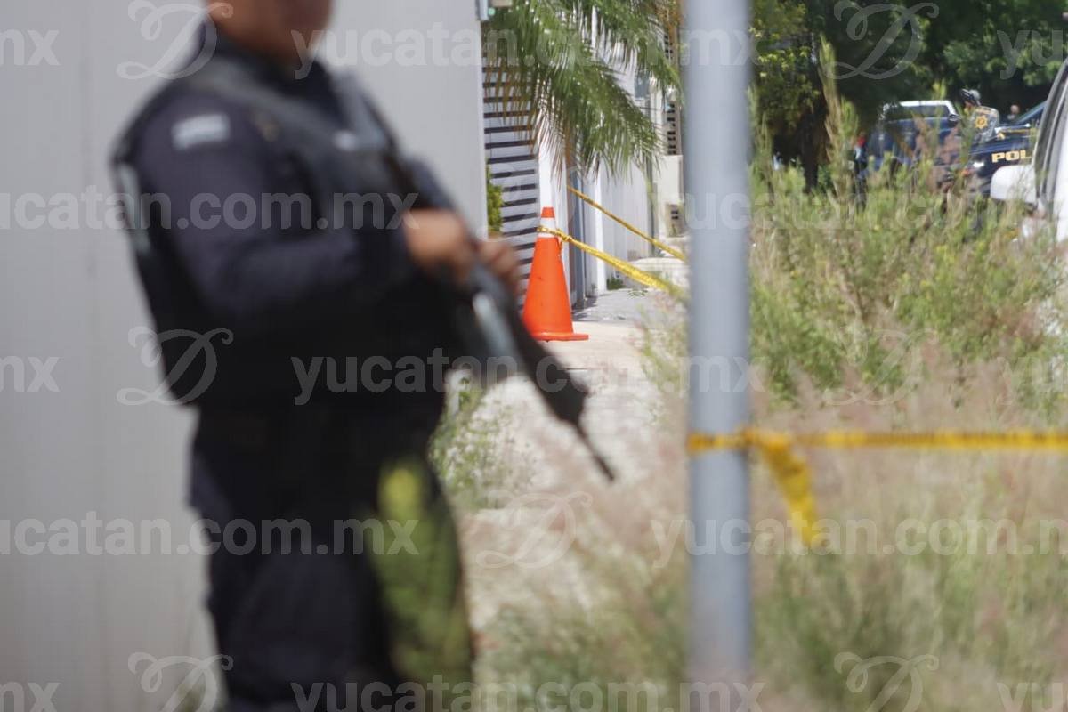 An armed officer in a black SSP uniform walks along yellow tape strung across a residential street next to a gray SUV in Mérida.