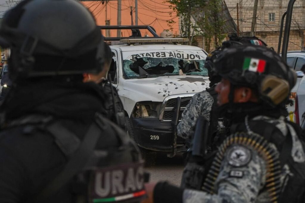 Mexican security forces in helmets stand in front of a white state police pickup with a shattered windshield and bullet holes