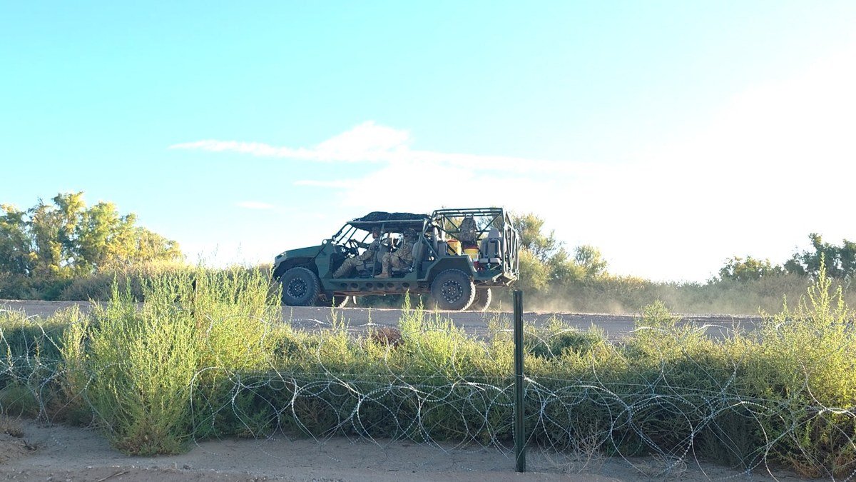 Uniformed troops ride an open military utility vehicle along a dirt levee road behind coils of concertina wire and tall brush