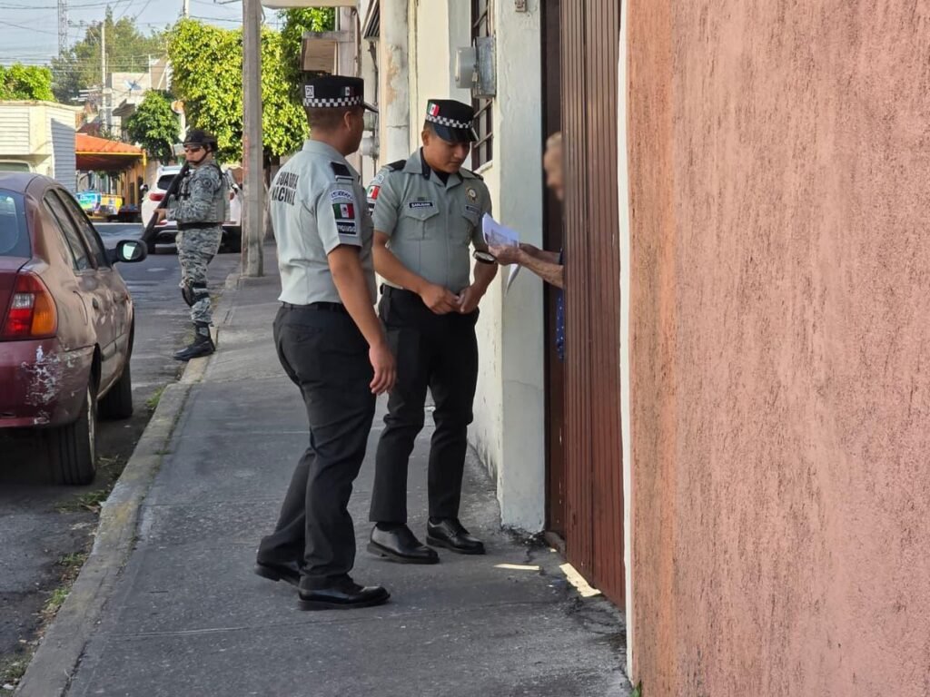Two Guardia Nacional officers speak with a resident at a doorway on a Morelos street while a soldier stands guard by a parked car.