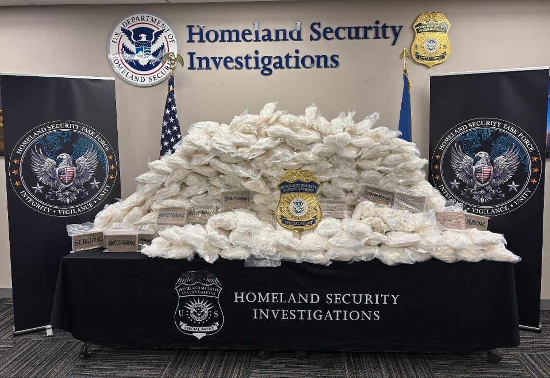Hundreds of clear plastic packages and several wrapped bricks piled on a black table under a Homeland Security Investigations sign, with an HSI badge placed in front
