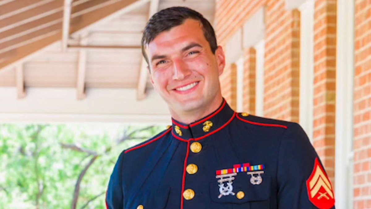 Nick Quets, a smiling young man with short dark hair, wearing a U.S. Marine Corps dress blue uniform with corporal chevrons and marksmanship badges, on a brick building's porch.