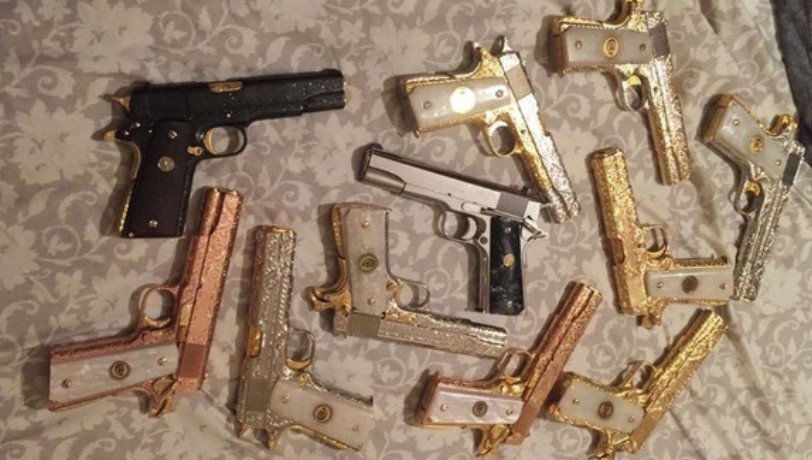 About a dozen ornate gold-, silver- and copper-colored pistols laid out on a patterned cloth