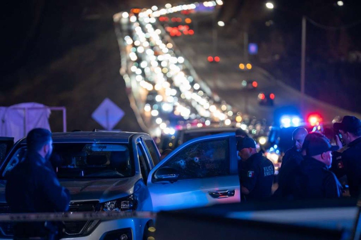 Municipal police officers stand beside a gray pickup with bullet holes in its passenger window at night on the road to Playas de Tijuana, with traffic backed up behind