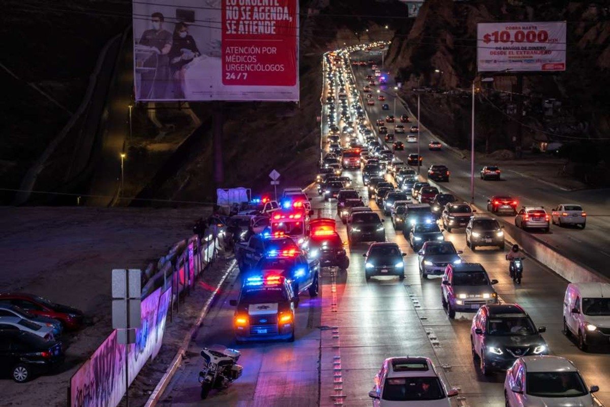 Police vehicles with flashing lights line the side of a busy highway at dusk in Tijuana as traffic backs up toward Playas de Tijuana
