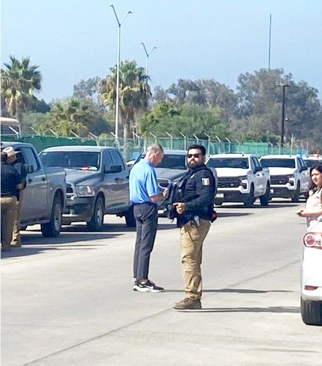 A man in a blue shirt stands beside an armed Mexican agent in a tactical vest next to a line of pickup trucks and SUVs on a road in Baja California