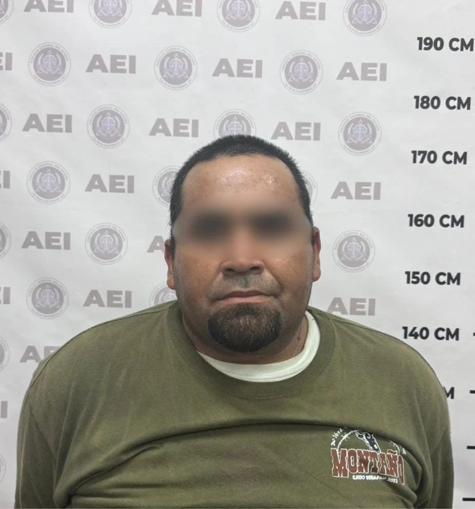 Booking photo with blurred face of a heavyset man in an olive T-shirt in front of a Baja California state investigations agency backdrop