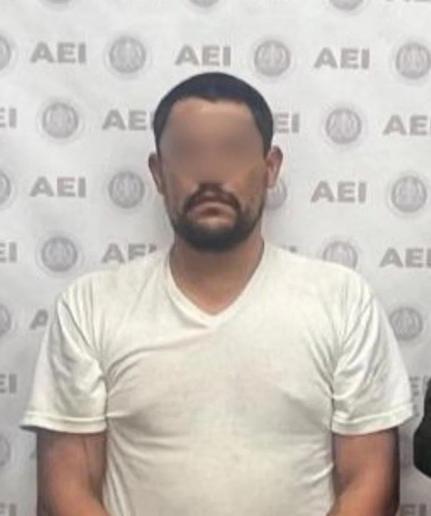 Booking photo with blurred face of a bearded man in a white T-shirt in front of a Baja California state investigations agency backdrop