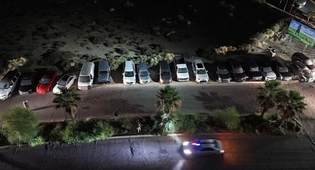 Night view from above of a row of cars and SUVs parked beside a desert lot in Puerto Peñasco, Sonora, with a police vehicle passing in the foreground