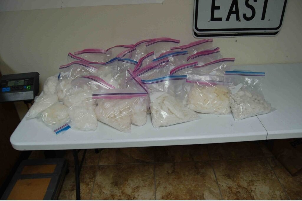 About two dozen zip-top bags of crystal methamphetamine piled on a white folding table beside a digital scale, under an 'East' road sign