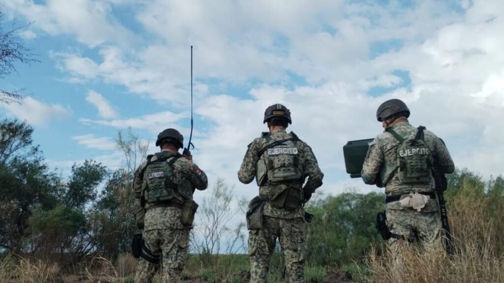 Three Mexican Army soldiers, one holding a radio antenna and one a portable device, stand on brush-covered ground in Tamaulipas