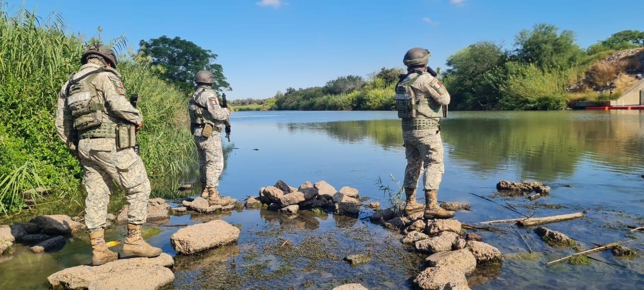 Three armed Mexican Army soldiers stand on rocks at the edge of the Rio Grande (Río Bravo) in Tamaulipas