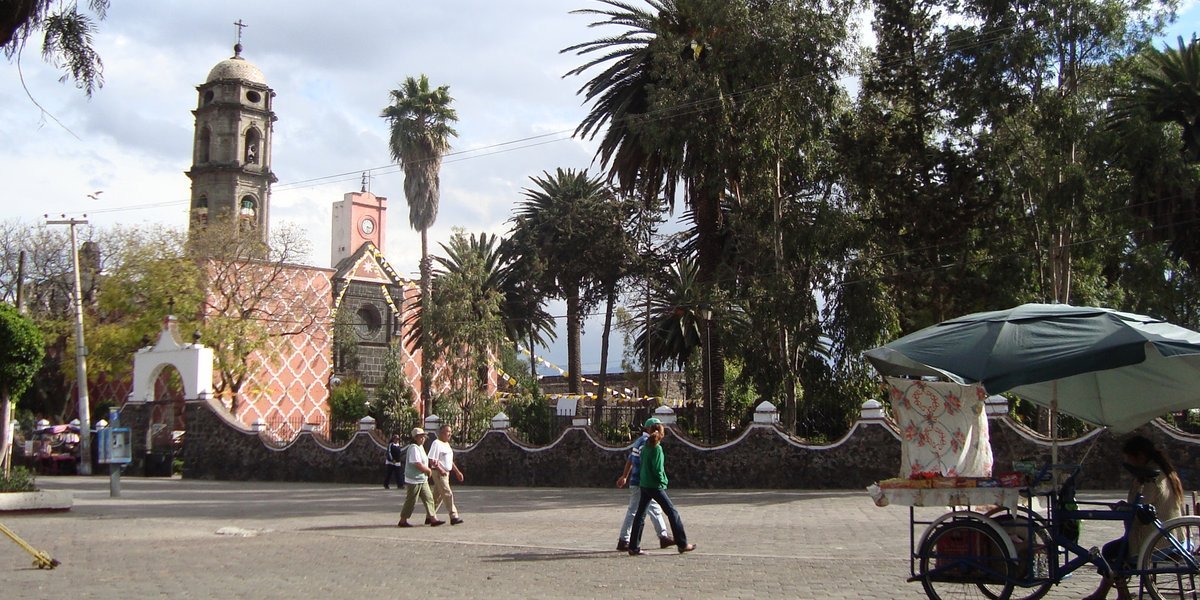 The central plaza of Tláhuac in Mexico City, with a colonial church bell tower, palm trees, a stone wall and people walking past a street vendor's tricycle cart.