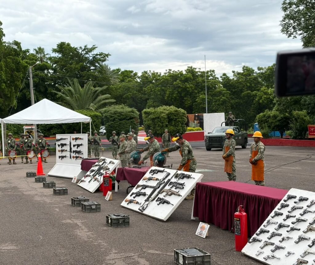 Mexican Army soldiers in orange welding aprons cut seized rifles with power saws beside display boards of confiscated rifles and pistols at a military base in Culiacán