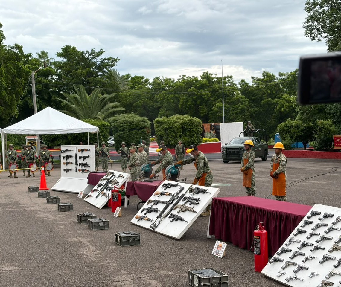 Mexican Army soldiers in orange welding aprons cut seized rifles with power saws beside display boards of confiscated rifles and pistols at a military base in Culiacán