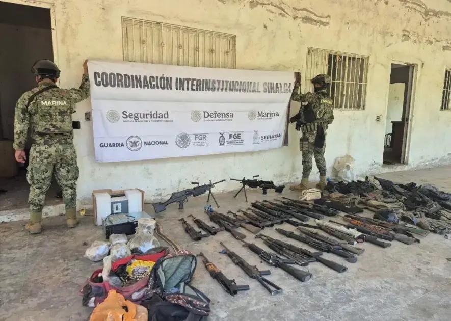 Two Mexican marines hang a 'Coordinación Interinstitucional Sinaloa' banner above rows of seized rifles, a belt-fed machine gun, magazines and bags laid out on a concrete floor