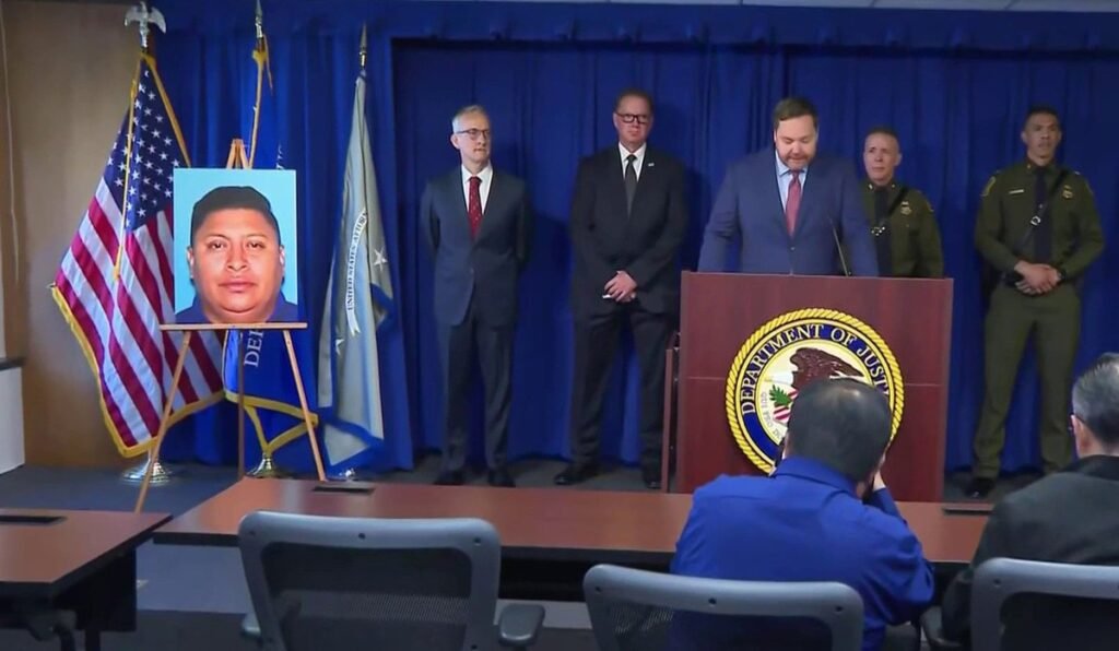 Federal officials in suits and Border Patrol uniforms stand behind a Department of Justice podium in Los Angeles, next to a man's photo displayed on an easel