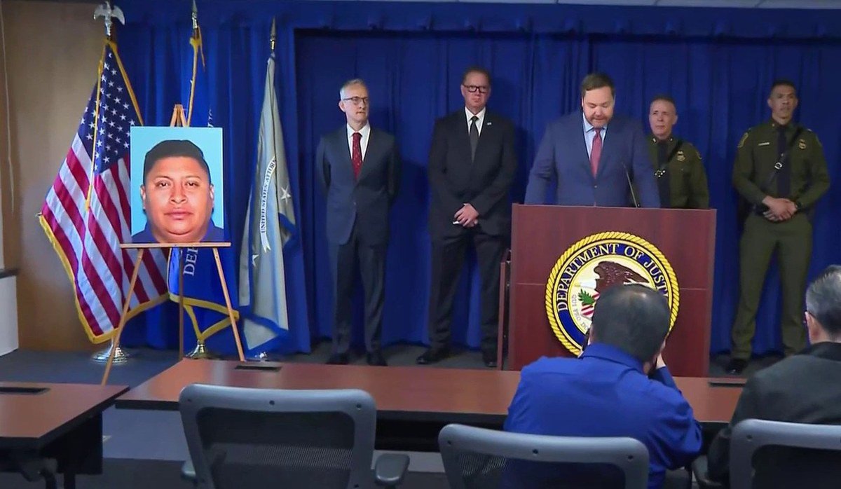 Federal officials in suits and Border Patrol uniforms stand behind a Department of Justice podium in Los Angeles, next to a man's photo displayed on an easel